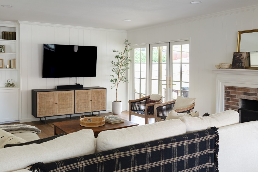 На фото: открытая гостиная комната среднего размера в стиле неоклассика (современная классика) с белыми стенами, полом из винила, стандартным камином, фасадом камина из кирпича, телевизором на стене и коричневым полом