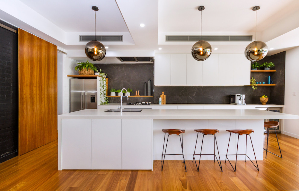 This is an example of an expansive contemporary galley open plan kitchen in Brisbane with a drop-in sink, flat-panel cabinets, quartz benchtops, black splashback, marble splashback, stainless steel appliances, medium hardwood floors, brown floor, white benchtop and with island.