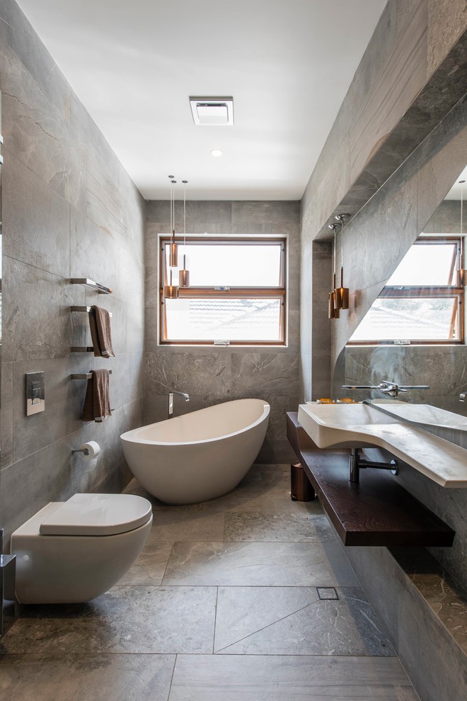 Contemporary master bathroom in Melbourne with open cabinets, a freestanding tub, a wall-mount toilet, gray tile, grey walls, an integrated sink and grey floor.