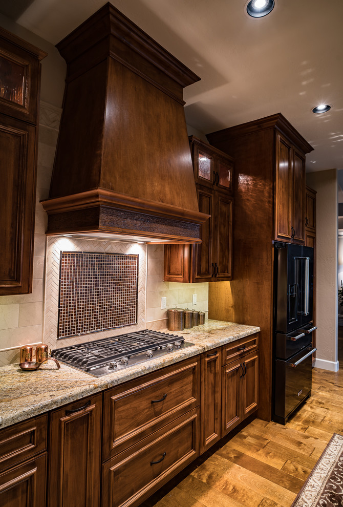 Mid-sized mountain style l-shaped medium tone wood floor and brown floor open concept kitchen photo in Minneapolis with a single-bowl sink, raised-panel cabinets, medium tone wood cabinets, granite countertops, beige backsplash, stone tile backsplash, black appliances and an island