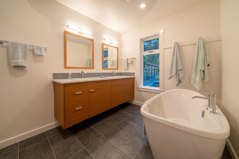 Inspiration for a beach style ensuite bathroom in Vancouver with flat-panel cabinets, brown cabinets, a freestanding bath, blue tiles, glass tiles, white walls, porcelain flooring, engineered stone worktops, grey floors, white worktops, double sinks and a floating vanity unit.