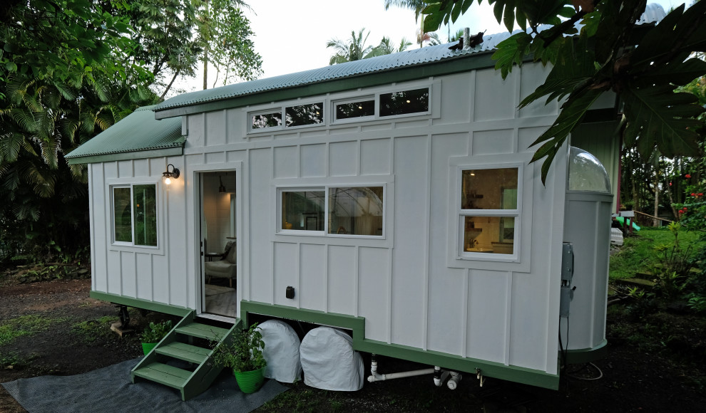 Small and white coastal two floor tiny house in Hawaii with wood cladding, a pitched roof, a metal roof, a grey roof and board and batten cladding.