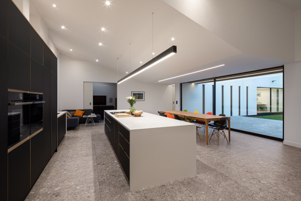 This is an example of a large modern galley open plan kitchen in Surrey with with island, grey floor, an integrated sink, flat-panel cabinets, black cabinets, quartzite benchtops, black appliances, ceramic floors, white benchtop and vaulted.