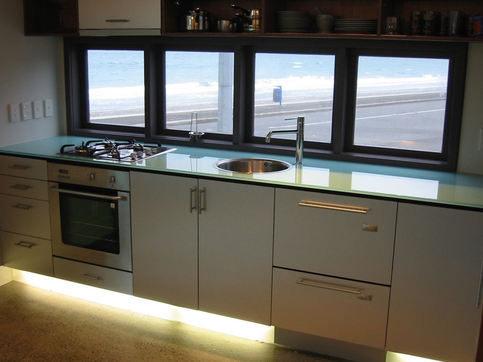 Inspiration for a small beach style l-shaped eat-in kitchen in Wellington with a single-bowl sink, flat-panel cabinets, stainless steel cabinets, glass benchtops, stainless steel appliances, concrete floors and no island.