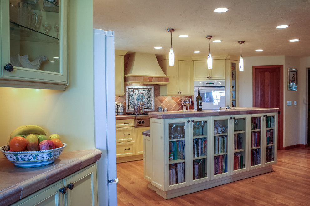 Photo of a large mediterranean l-shaped eat-in kitchen in Seattle with recessed-panel cabinets, yellow cabinets, tile benchtops, orange splashback, ceramic splashback, with island, white appliances and light hardwood floors.