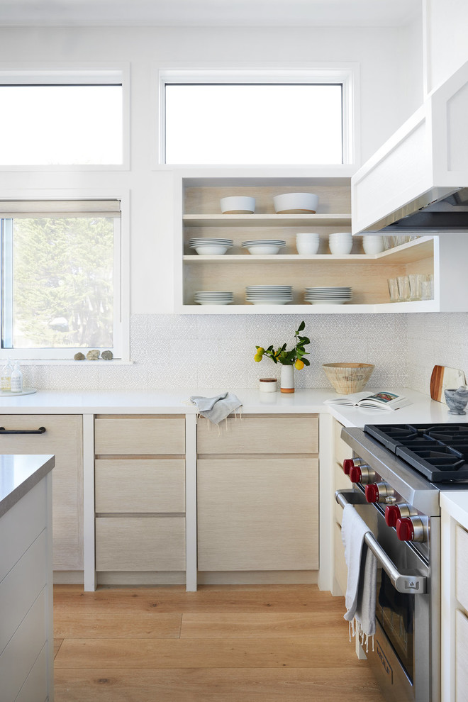 Inspiration for a beach style l-shaped kitchen in San Francisco with flat-panel cabinets, light wood cabinets, white splashback, stainless steel appliances, medium hardwood floors, with island, brown floor and white benchtop.