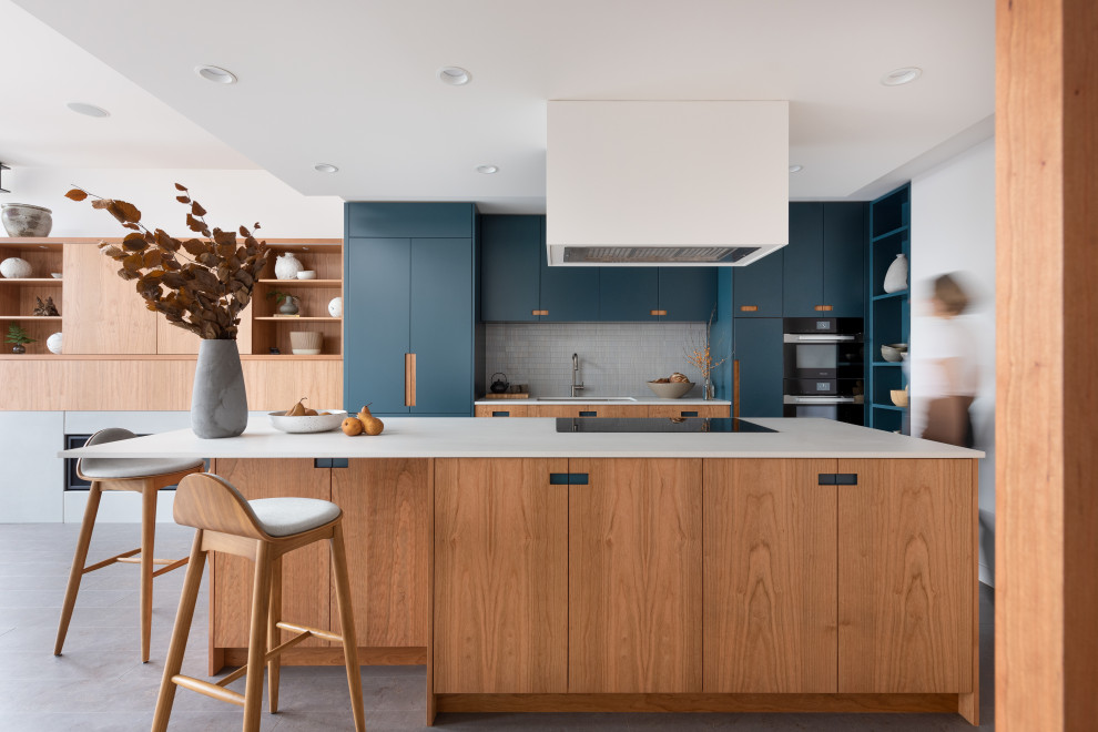 This is an example of a small contemporary l-shaped open plan kitchen in Vancouver with an undermount sink, flat-panel cabinets, blue cabinets, grey splashback, stone tile splashback, black appliances, cork floors, with island, grey floor and white benchtop.