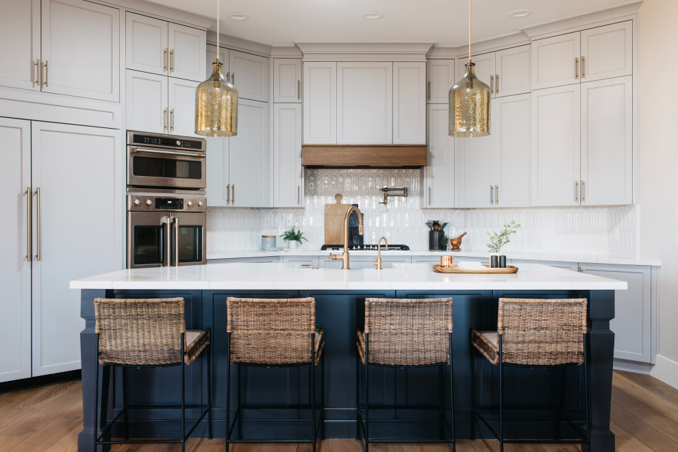 Mid-sized beach style eat-in kitchen in Phoenix with a farmhouse sink, recessed-panel cabinets, blue cabinets, marble benchtops, white splashback, stone tile splashback, panelled appliances, medium hardwood floors, with island, brown floor and white benchtop.