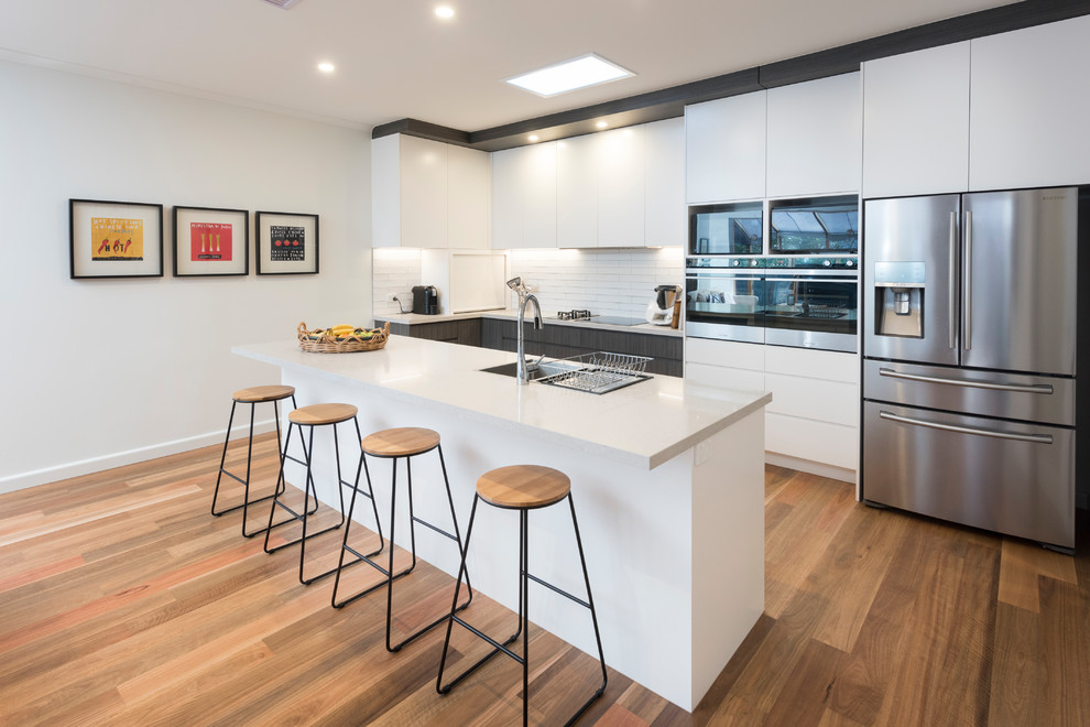 This is an example of a contemporary single-wall eat-in kitchen in Melbourne with an undermount sink, flat-panel cabinets, white cabinets, white splashback, white appliances, medium hardwood floors, with island, brown floor, granite benchtops and subway tile splashback.