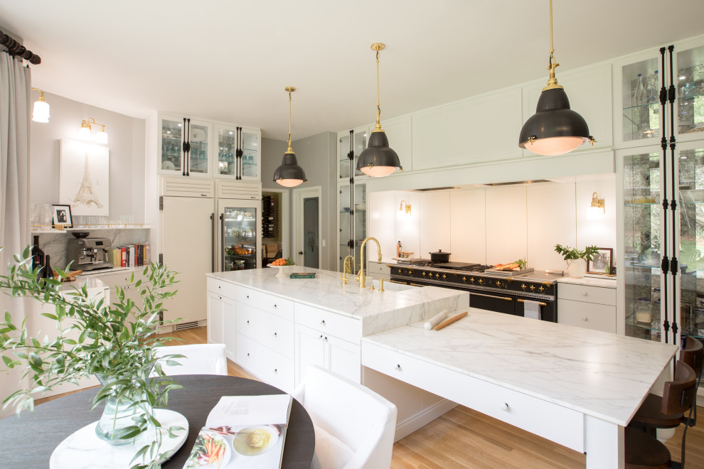 This is an example of a medium sized l-shaped kitchen/diner in Seattle with a belfast sink, glass-front cabinets, yellow cabinets, marble worktops, white splashback, porcelain splashback, white appliances, medium hardwood flooring, an island, brown floors and white worktops.