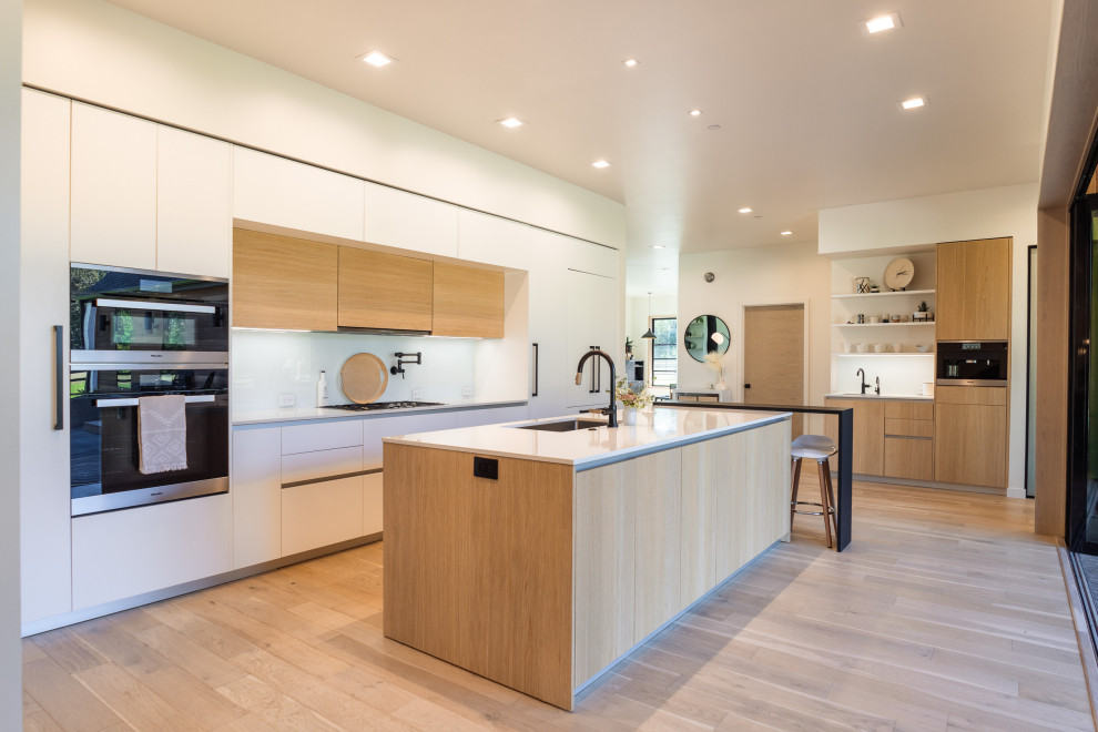 This is an example of an expansive contemporary single-wall eat-in kitchen in Portland with an undermount sink, flat-panel cabinets, light wood cabinets, quartz benchtops, white splashback, glass sheet splashback, panelled appliances, light hardwood floors, with island, brown floor and white benchtop.