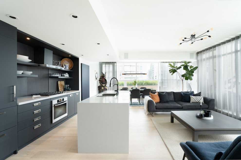 Large contemporary galley open plan kitchen in Vancouver with flat-panel cabinets, black cabinets, black splashback, stainless steel appliances, with island, beige floor, white benchtop, an undermount sink, solid surface benchtops, slate splashback and light hardwood floors.