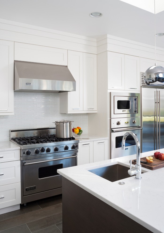 Example of a mid-sized transitional l-shaped ceramic tile and brown floor enclosed kitchen design in DC Metro with an undermount sink, shaker cabinets, white cabinets, quartz countertops, white backsplash, glass tile backsplash, stainless steel appliances and an island