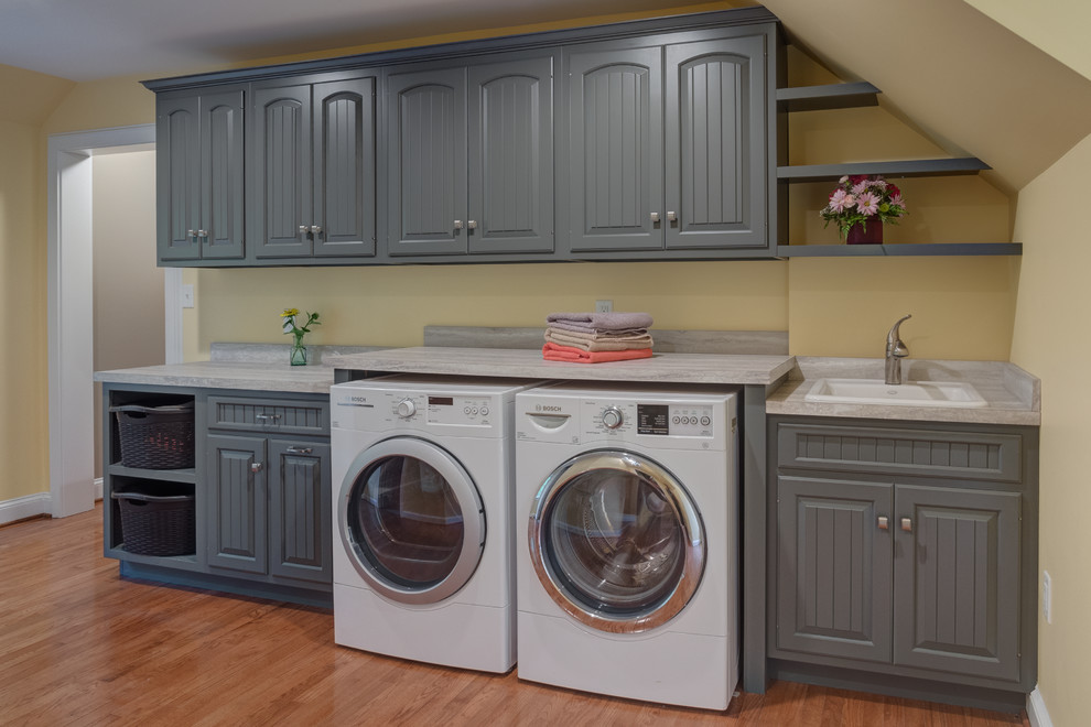 Photo of a mid-sized country galley dedicated laundry room in Other with a drop-in sink, raised-panel cabinets, grey cabinets, medium hardwood floors, a side-by-side washer and dryer and beige walls.