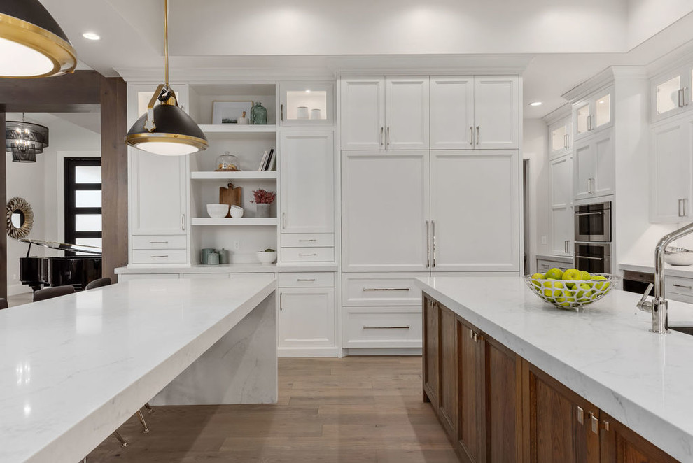 Photo of a large transitional l-shaped eat-in kitchen in Salt Lake City with an undermount sink, recessed-panel cabinets, white cabinets, quartzite benchtops, white splashback, marble splashback, panelled appliances, light hardwood floors, multiple islands, beige floor and white benchtop.