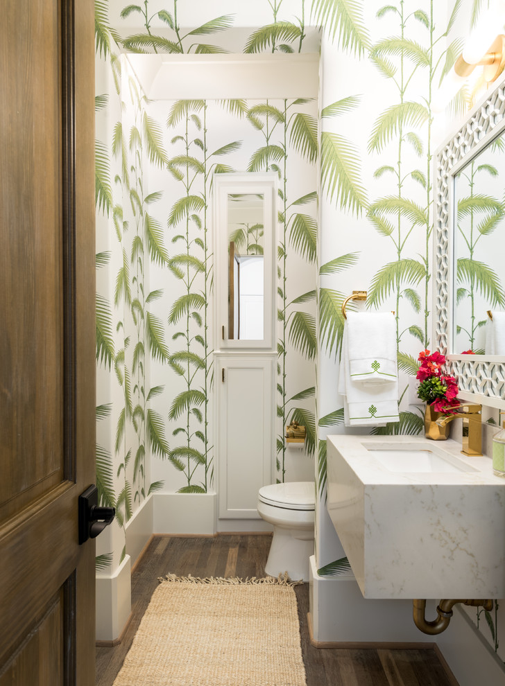 Photo of a tropical powder room in Dallas with white cabinets, a one-piece toilet, multi-coloured walls, medium hardwood floors, engineered quartz benchtops, brown floor and an undermount sink.
