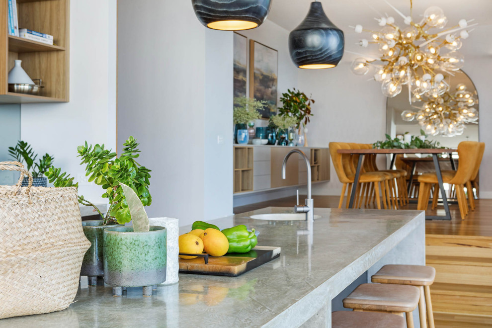 Inspiration for a mid-sized beach style galley kitchen in Sydney with a drop-in sink, flat-panel cabinets, black cabinets, concrete benchtops, metallic splashback, mirror splashback, light hardwood floors, with island and grey benchtop.