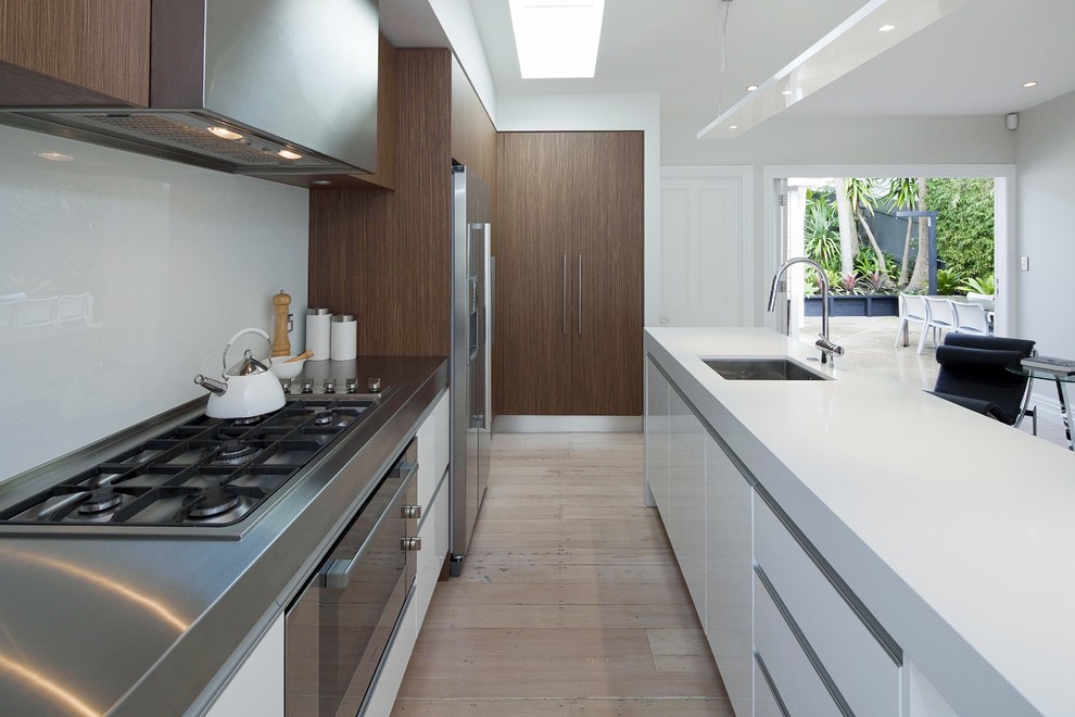 Inspiration for a small contemporary galley kitchen in Auckland with stainless steel appliances, stainless steel benchtops, flat-panel cabinets, white cabinets and white splashback.