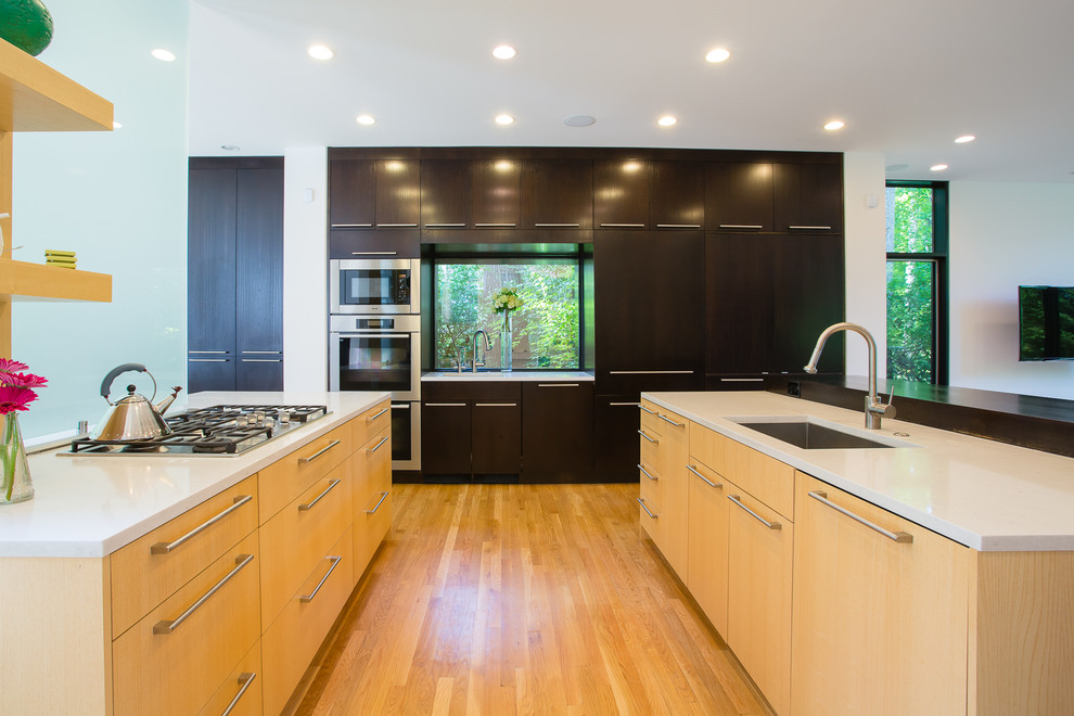 This is an example of a mid-sized contemporary u-shaped open plan kitchen in DC Metro with flat-panel cabinets, light wood cabinets, stainless steel appliances, an undermount sink, solid surface benchtops, light hardwood floors, multiple islands, beige floor and white benchtop.