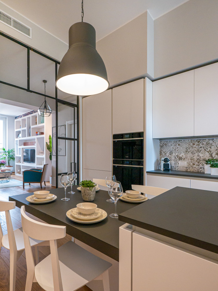 Large contemporary galley separate kitchen in Milan with an integrated sink, flat-panel cabinets, white cabinets, solid surface benchtops, multi-coloured splashback, porcelain splashback, black appliances, light hardwood floors, with island, grey benchtop and recessed.