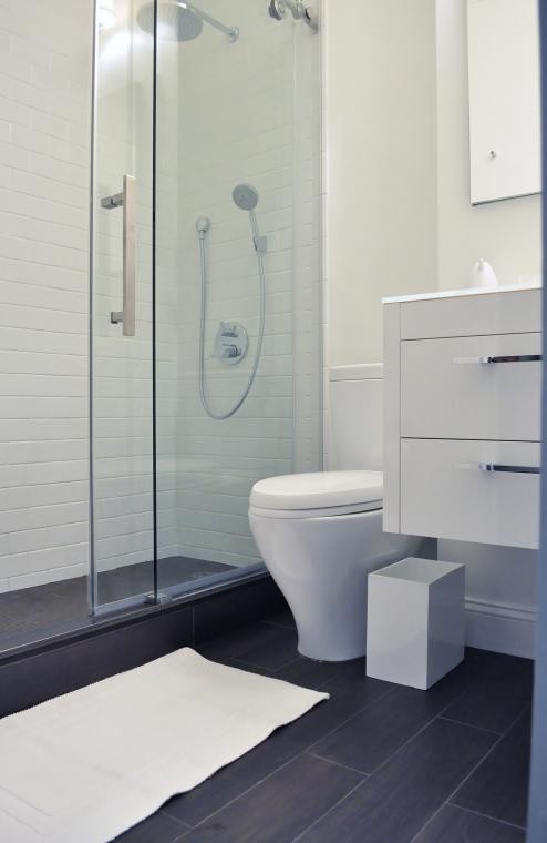 This is an example of a small modern 3/4 bathroom in New York with a wall-mount sink, flat-panel cabinets, white cabinets, a corner shower, white tile, subway tile, white walls, a two-piece toilet and vinyl floors.
