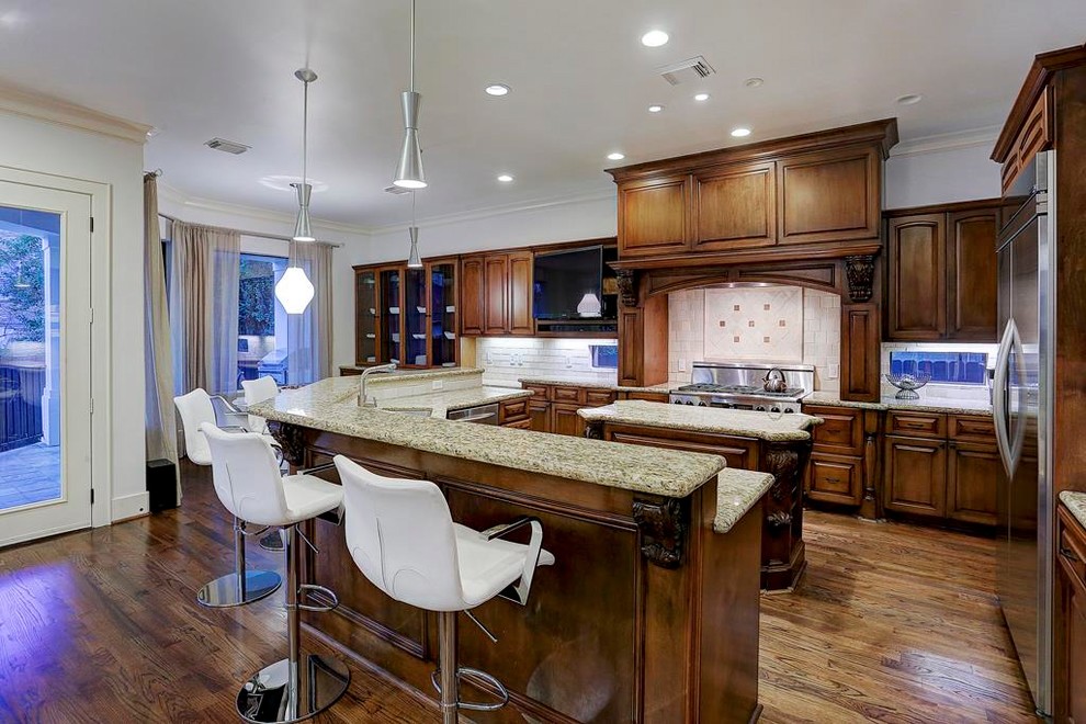 This is an example of a large mediterranean u-shaped open plan kitchen in Houston with a double-bowl sink, raised-panel cabinets, granite benchtops, white splashback, travertine splashback, stainless steel appliances, medium hardwood floors, multiple islands, brown floor, beige benchtop and dark wood cabinets.