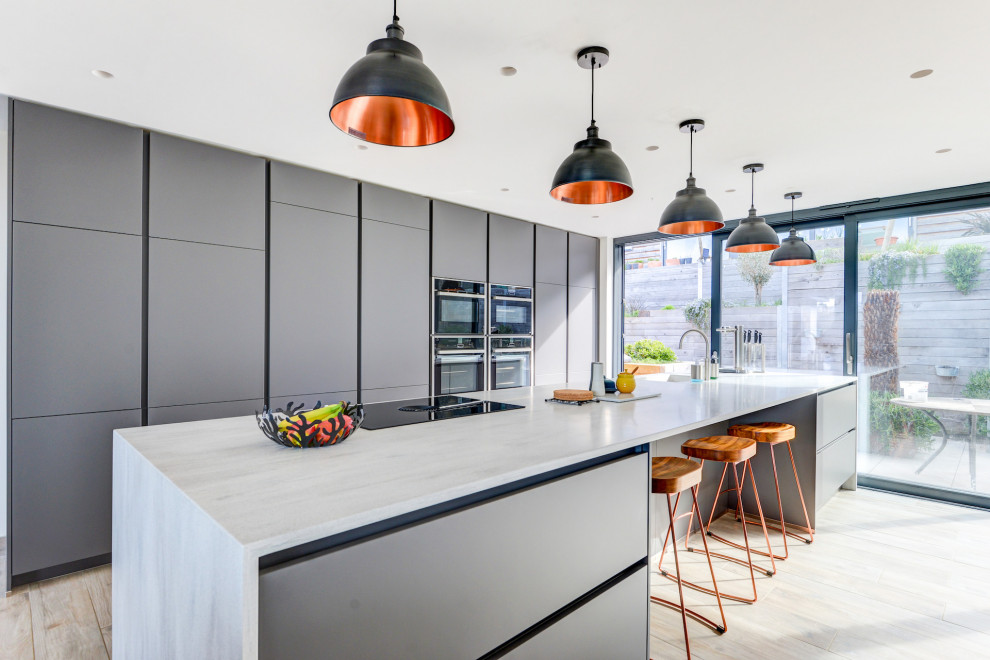 Inspiration for an expansive modern single-wall open plan kitchen in Sussex with an integrated sink, flat-panel cabinets, grey cabinets, solid surface benchtops, black appliances, light hardwood floors, with island, brown floor and grey benchtop.