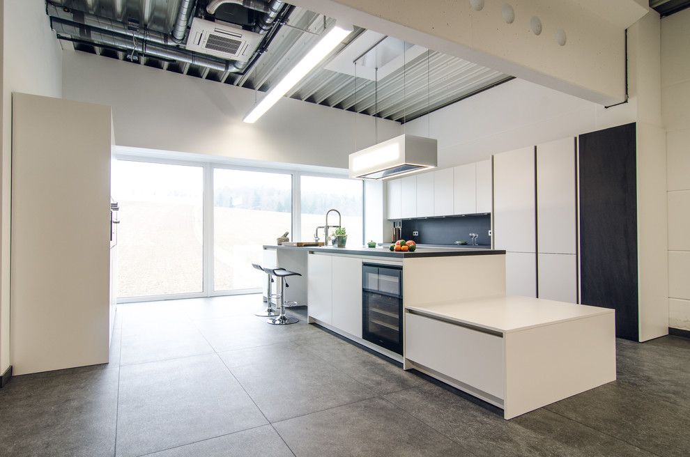This is an example of an expansive industrial galley open plan kitchen in Frankfurt with with island, flat-panel cabinets, white cabinets, black splashback, grey floor, grey benchtop and black appliances.