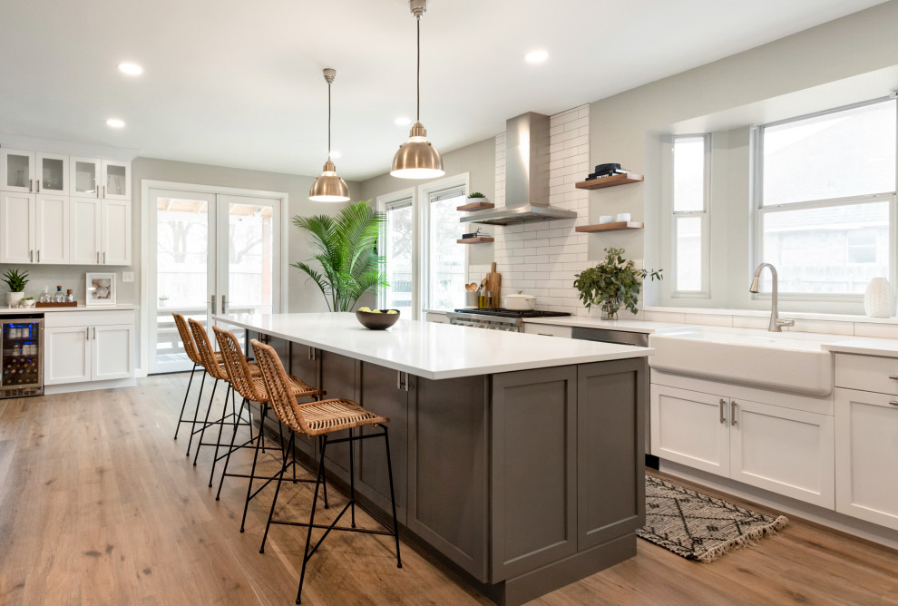 Example of a transitional galley medium tone wood floor and brown floor kitchen design in Austin with a farmhouse sink, shaker cabinets, white cabinets, white backsplash, stainless steel appliances, an island and white countertops