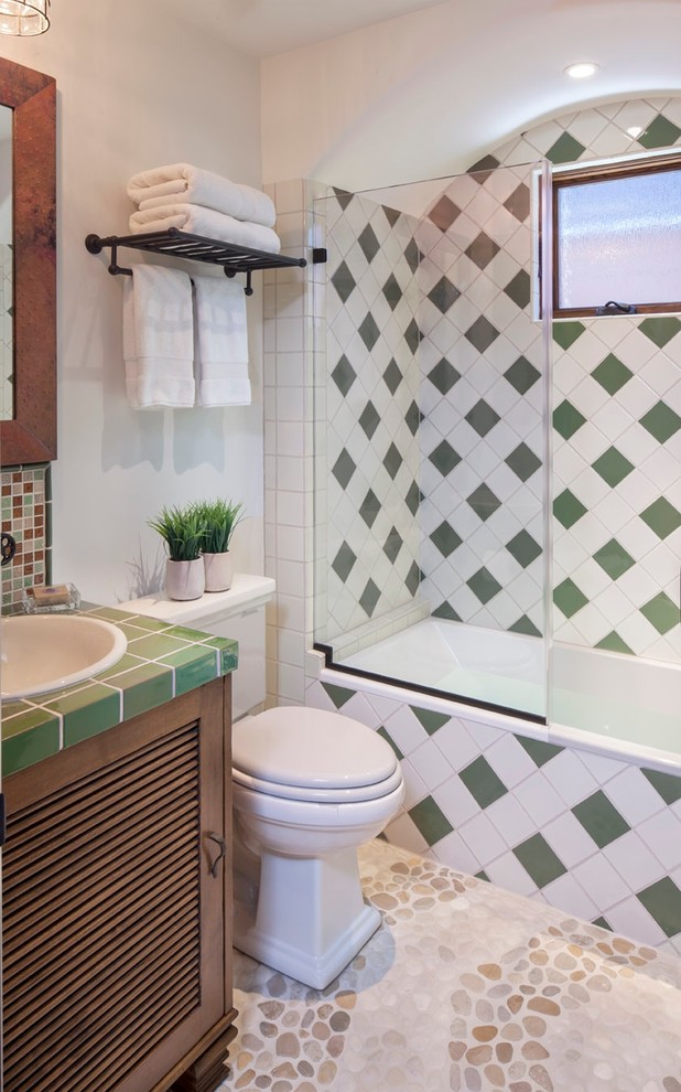 This is an example of a mediterranean bathroom in Los Angeles with a drop-in sink, medium wood cabinets, tile benchtops, an alcove tub, a shower/bathtub combo, a two-piece toilet, multi-coloured tile, white walls and pebble tile floors.