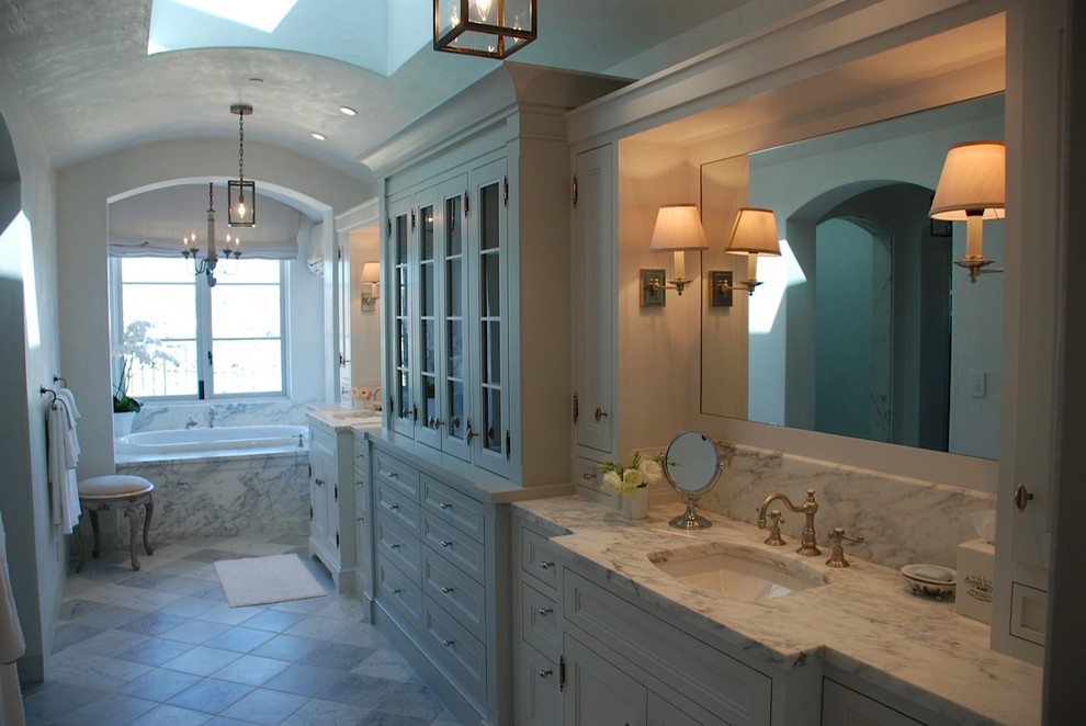 This is an example of a mid-sized mediterranean master bathroom in Orange County with beaded inset cabinets, white cabinets, a drop-in tub, limestone floors, an undermount sink, marble benchtops and multi-coloured floor.