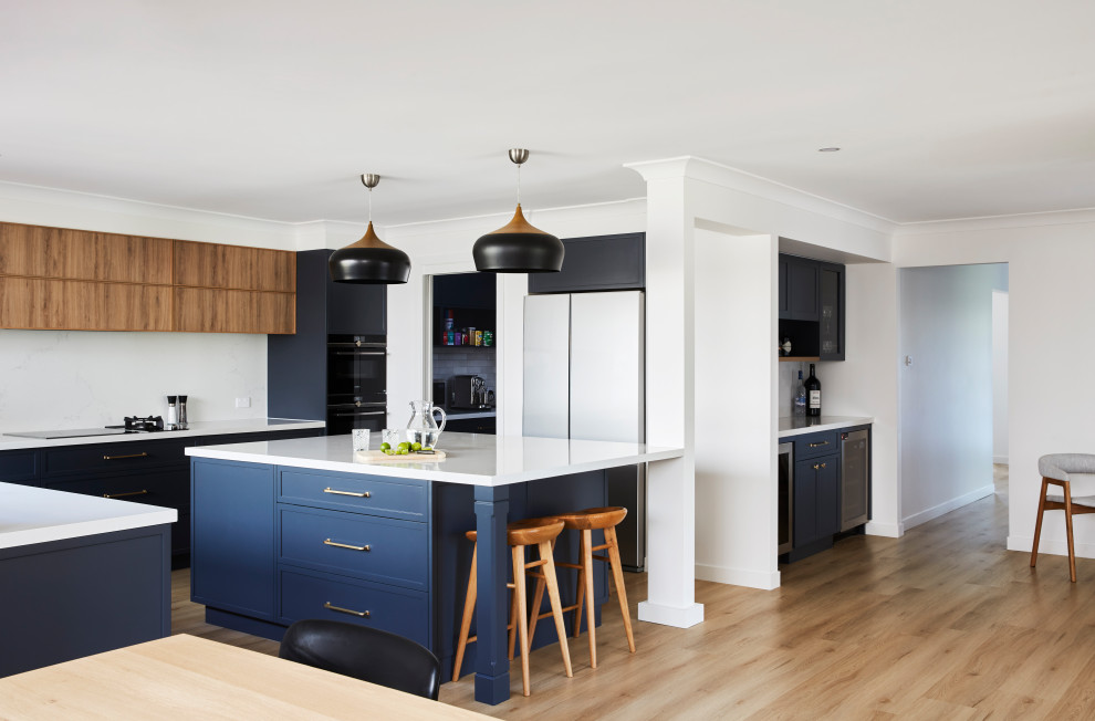 Inspiration for an expansive transitional l-shaped open plan kitchen in Sydney with a double-bowl sink, shaker cabinets, blue cabinets, quartz benchtops, white splashback, engineered quartz splashback, black appliances, laminate floors, with island, brown floor and white benchtop.