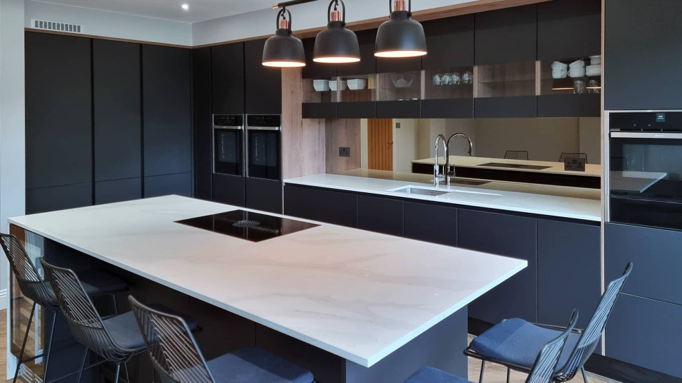 Example of a large minimalist single-wall light wood floor and multicolored floor open concept kitchen design in Hertfordshire with an integrated sink, flat-panel cabinets, black cabinets, quartzite countertops, mirror backsplash, black appliances, an island and white countertops