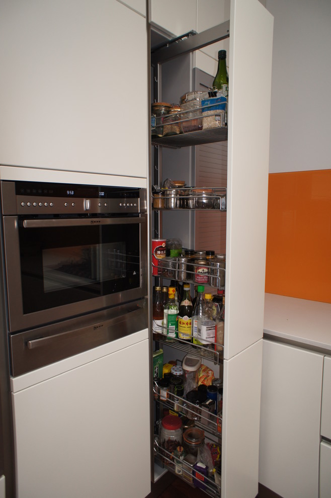 This is an example of a mid-sized modern u-shaped open plan kitchen in London with a single-bowl sink, flat-panel cabinets, solid surface benchtops, orange splashback, glass sheet splashback, stainless steel appliances, no island and white cabinets.