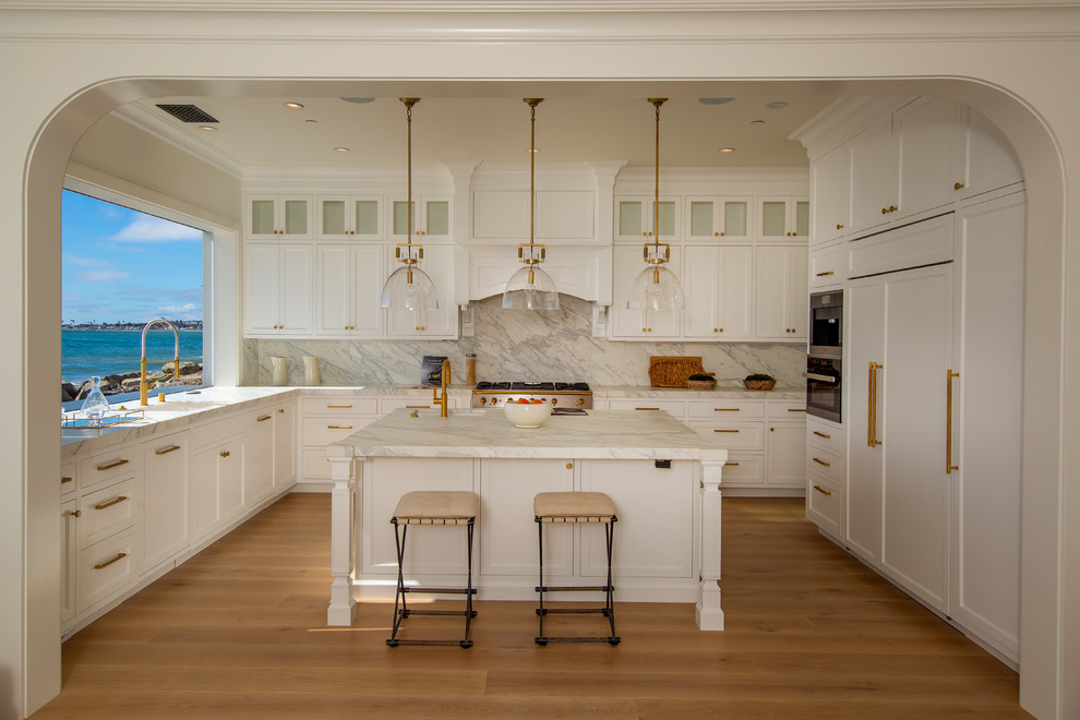 Beach style u-shaped kitchen in Los Angeles with an integrated sink, recessed-panel cabinets, white cabinets, white splashback, window splashback, panelled appliances, medium hardwood floors, with island, brown floor and white benchtop.
