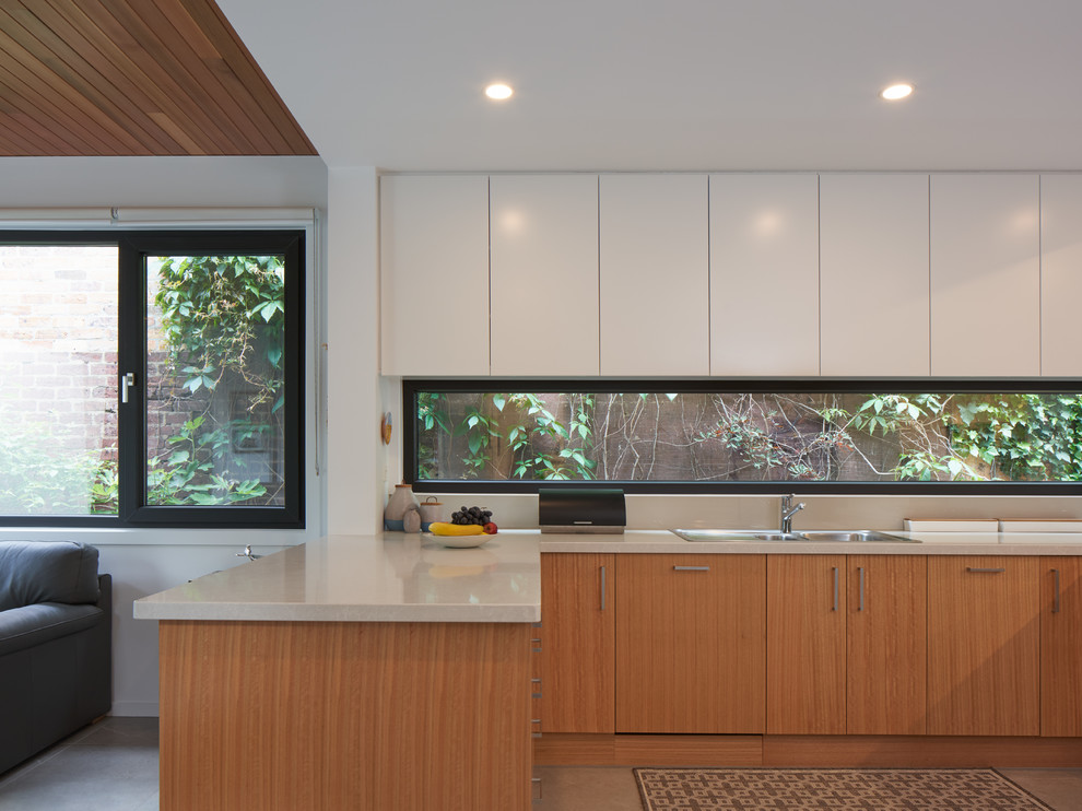 This is an example of a mid-sized contemporary u-shaped kitchen in Melbourne with a drop-in sink, flat-panel cabinets, medium wood cabinets, window splashback, panelled appliances, a peninsula, beige floor and beige benchtop.