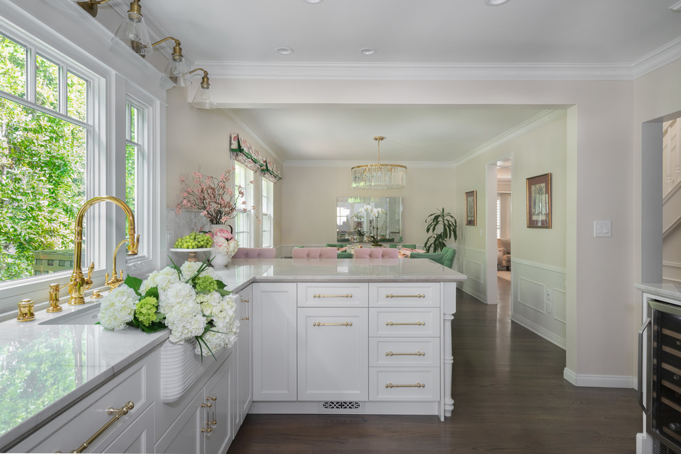 Mid-sized traditional galley eat-in kitchen in Los Angeles with a farmhouse sink, recessed-panel cabinets, white cabinets, quartzite benchtops, white splashback, marble splashback, white appliances, medium hardwood floors, a peninsula, brown floor and white benchtop.