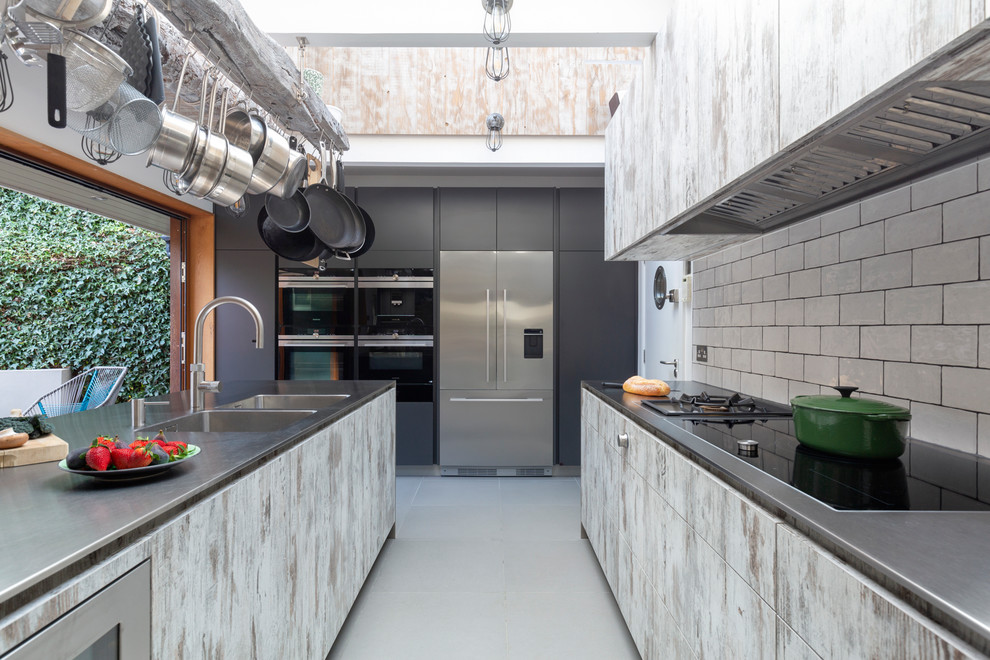 Photo of a large industrial u-shaped kitchen in London with an integrated sink, flat-panel cabinets, stainless steel benchtops, white splashback, stainless steel appliances, grey floor, grey cabinets, subway tile splashback and grey benchtop.