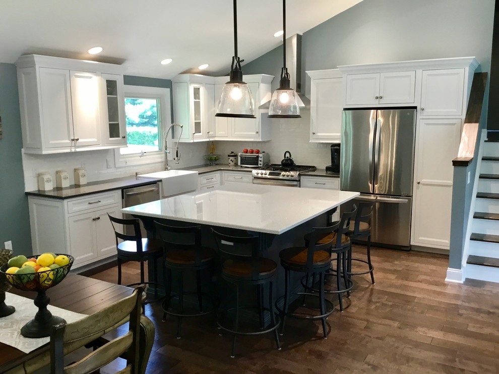Example of a mid-sized transitional l-shaped dark wood floor and brown floor kitchen design in New York with an island, a farmhouse sink, recessed-panel cabinets, white cabinets, granite countertops, white backsplash, subway tile backsplash, stainless steel appliances and black countertops