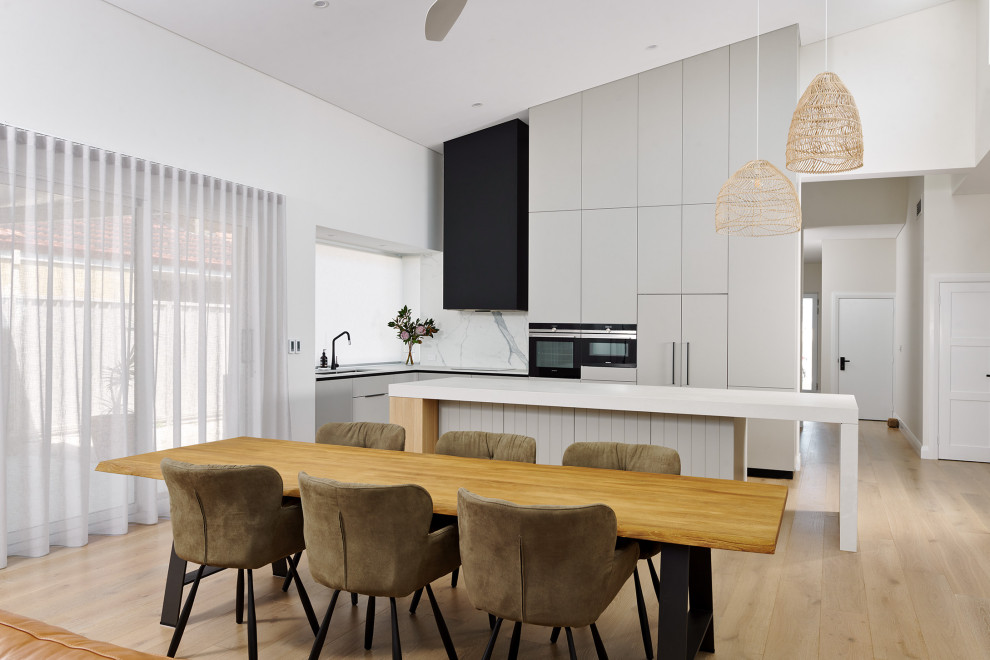 This is an example of a large scandinavian l-shaped kitchen/diner in Perth with a double-bowl sink, flat-panel cabinets, grey cabinets, engineered stone countertops, white splashback, porcelain splashback, black appliances, light hardwood flooring, an island, beige floors, white worktops and a vaulted ceiling.