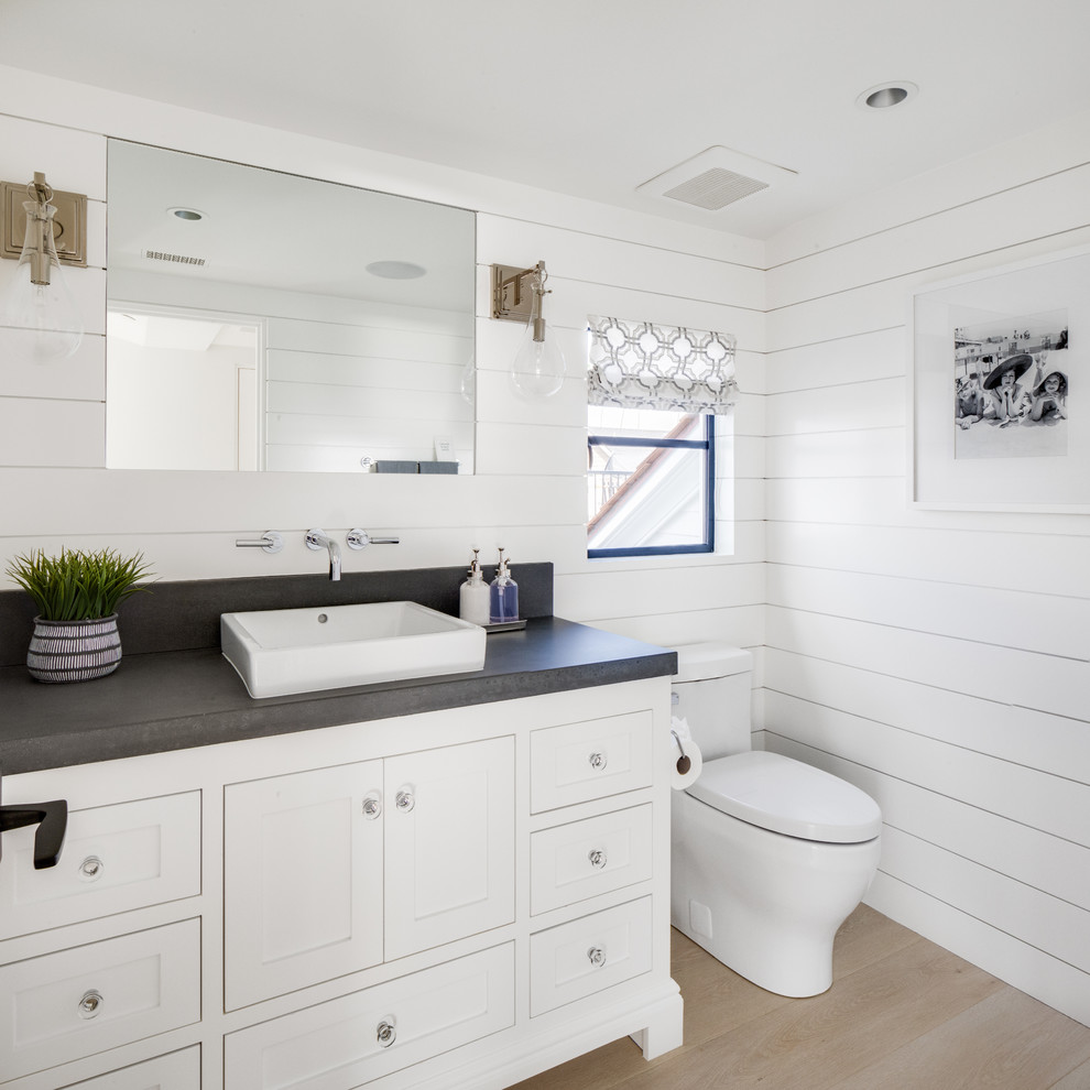 Photo of a beach style powder room in Orange County with furniture-like cabinets, white cabinets, a one-piece toilet, white walls, light hardwood floors, a vessel sink, beige floor and grey benchtops.