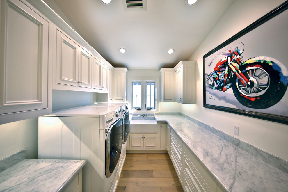 Design ideas for a large traditional galley dedicated laundry room in Orange County with a farmhouse sink, beaded inset cabinets, white cabinets, marble benchtops, light hardwood floors and a side-by-side washer and dryer.