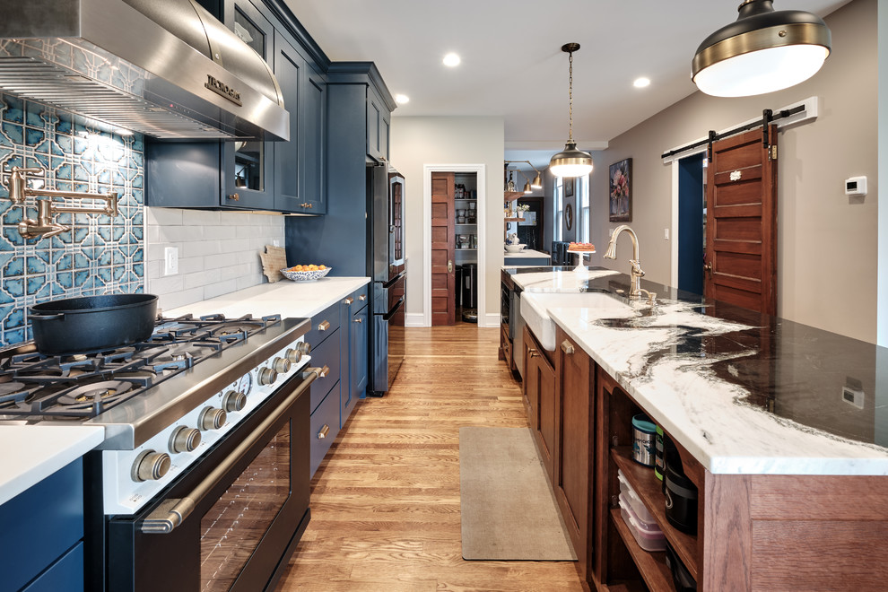 Large eclectic galley open plan kitchen in Philadelphia with a farmhouse sink, recessed-panel cabinets, blue cabinets, quartzite benchtops, grey splashback, ceramic splashback, black appliances, medium hardwood floors, with island, brown floor and multi-coloured benchtop.