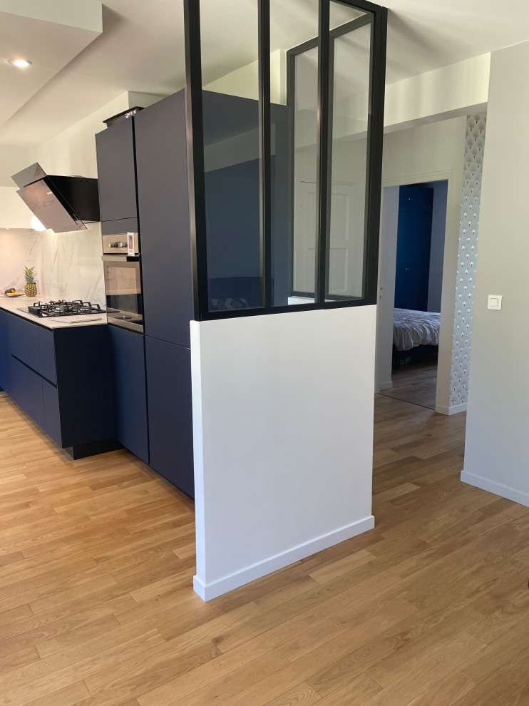 Mid-sized industrial u-shaped kitchen in Paris with flat-panel cabinets, blue cabinets, white splashback, black appliances, light hardwood floors, no island, brown floor and white benchtop.