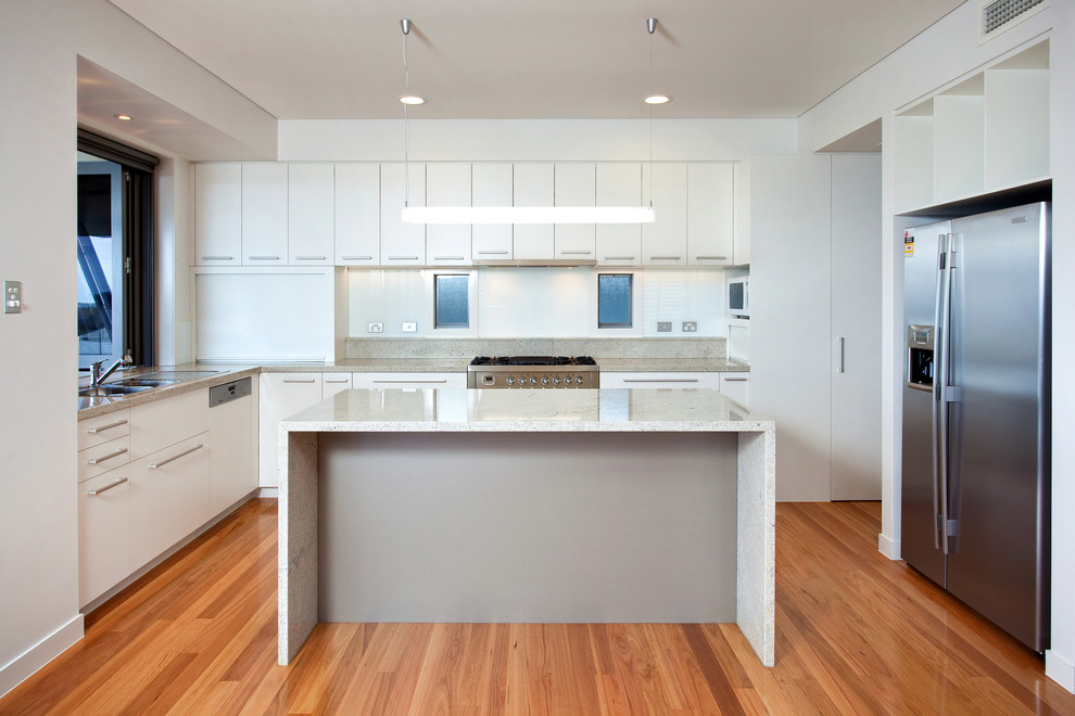 Inspiration for a large contemporary l-shaped open plan kitchen in Brisbane with a drop-in sink, white splashback, glass sheet splashback, stainless steel appliances, with island, flat-panel cabinets, white cabinets, light hardwood floors and brown floor.