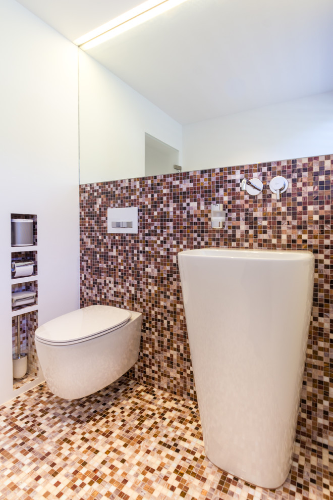 This is an example of a small contemporary powder room in Stuttgart with open cabinets, white cabinets, a wall-mount toilet, beige tile, brown tile, orange tile, red tile, black tile, mosaic tile, white walls, mosaic tile floors, a pedestal sink and multi-coloured floor.