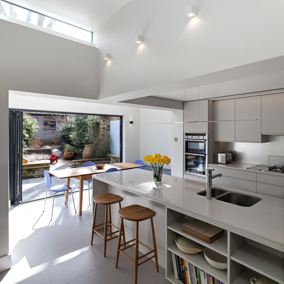 This is an example of a contemporary eat-in kitchen in London with a double-bowl sink, flat-panel cabinets, grey cabinets, white splashback, glass sheet splashback and with island.