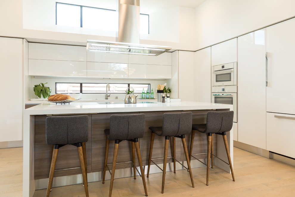 Photo of a large contemporary l-shaped open plan kitchen in San Francisco with an undermount sink, flat-panel cabinets, white cabinets, quartz benchtops, white splashback, white appliances, light hardwood floors, with island and beige floor.