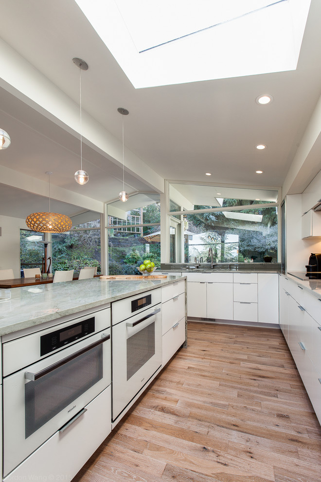 Photo of a large contemporary l-shaped eat-in kitchen in Seattle with an undermount sink, flat-panel cabinets, white cabinets, quartzite benchtops, grey splashback, glass tile splashback, white appliances, light hardwood floors, with island and beige floor.