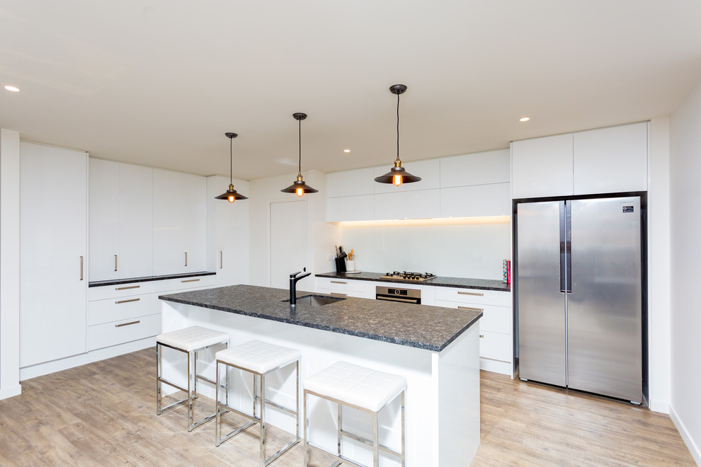 This is an example of a mid-sized modern u-shaped open plan kitchen in Christchurch with an undermount sink, flat-panel cabinets, white cabinets, granite benchtops, white splashback, glass sheet splashback, stainless steel appliances, light hardwood floors, with island, brown floor and black benchtop.